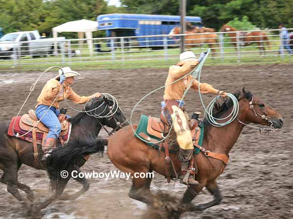 Two cowgirls gallop down a muddy arena